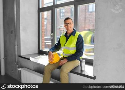 architecture, construction business and people concept - happy smiling male builder or architect with tumbler or thermo cup sitting on window sill at office. happy builder with tumbler sitting on window sill