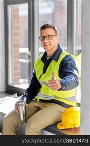 architecture, construction business and people concept - happy smiling male builder or architect with tumbler or thermo cup sitting on window sill and showing thumbs up at office. happy builder with tumbler showing thumbs up