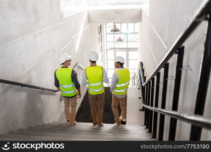 architecture, construction business and people concept - male architects in helmets with blueprints walking downstairs at office building. architects in helmets walking downstairs at office