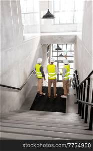 architecture, construction business and people concept - male architects in helmets with blueprints on stairs at office building. architects in helmets with blueprints at office