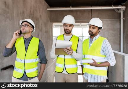architecture, construction business and people concept - male architects in helmets with smartphone, blueprint and clipboard working at office building. male architects in helmets working at office