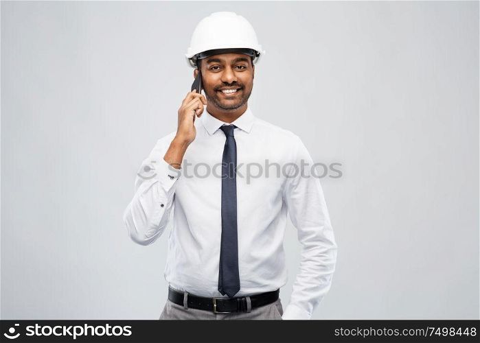 architecture, construction business and people concept - smiling indian male architect in helmet calling on smartphone over grey background. indian male architect calling on smartphone