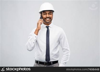 architecture, construction business and people concept - smiling indian male architect in helmet calling on smartphone over grey background. indian male architect calling on smartphone