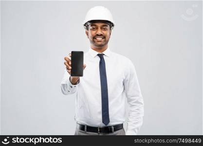 architecture, construction business and people concept - smiling indian male architect in helmet showing smartphone over grey background. indian male architect in helmet showing smartphone