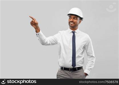 architecture, construction business and people concept - smiling indian male architect in helmet pointing finger to something over grey background. indian male architect in helmet showing something