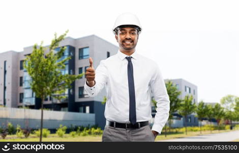 architecture, construction business and people concept - smiling indian male architect in helmet showing thumbs up over living houses on city street background. architect showing thumbs up on city street