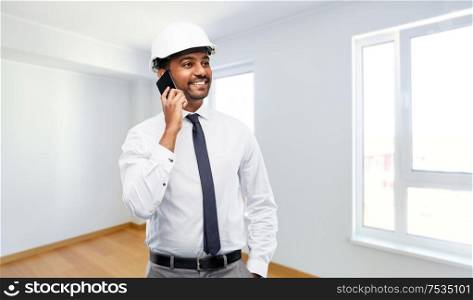 architecture, construction business and people concept - smiling indian male architect in helmet calling on smartphone over empty new apartment room background. indian male architect calling on smartphone
