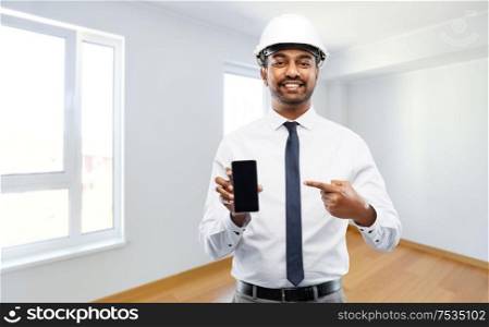 architecture, construction business and people concept - smiling indian male architect in helmet showing smartphone over empty new apartment room background. indian male architect in helmet showing smartphone
