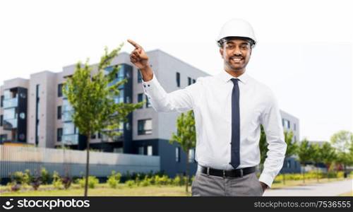 architecture, construction business and people concept - smiling indian male architect in helmet pointing finger to something over living houses on city street background. architect in helmet pointing finger on city street