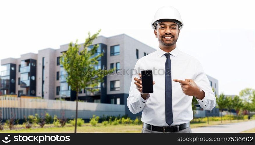 architecture, construction business and people concept - smiling indian male architect in helmet showing smartphone over living houses on city street background. architect showing smartphone on city street