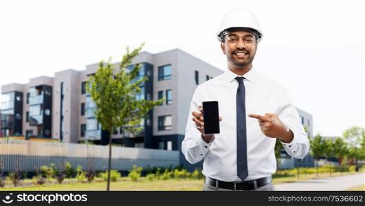 architecture, construction business and people concept - smiling indian male architect in helmet showing smartphone over living houses on city street background. architect showing smartphone on city street