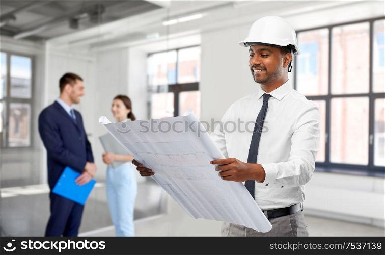 architecture, construction business and people concept - smiling indian male architect in helmet with blueprint over empty room of new office background. indian male architect in helmet with blueprint