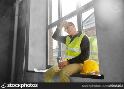 architecture, construction business and people concept - tired male builder or architect with tumbler or thermo cup sitting on window sill at office. tired builder with tumbler sitting on window sill