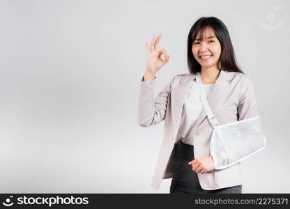 Arm broken. Business woman confident smile broken arm after accident wear arm splint for treatment show OK sign, happy Asian female sling support hand studio isolated on white background, copy space