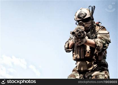 Army elite forces soldier, U.S. marine rider, special security guard in helmet and camo uniform, standing on background of clear blue sky, aiming assault rifle with optical sight at camera. Copyspace. Army special forces fighter aiming gun at camera