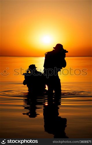 Army soldiers with rifles orange sunset silhouette in action during raid crossing river in the water. Army soldier silhouette