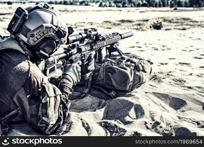 Army special forces soldier, US marine riders regiment fighter, private military company mercenary, lying on sand, aiming service rifle, supporting running or attacking comrade with firearm fire. Marine riders team attacking enemy in sandy area