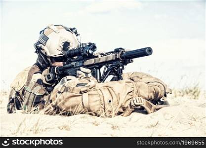 Army special forces soldier, US marine riders regiment fighter, private military company mercenary, lying on sand, aiming service rifle, supporting running or attacking comrade with firearm fire. Marine riders team attacking enemy in sandy area