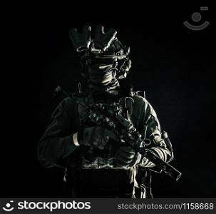Army special operations forces soldier in mask and combat uniform, helmet equipped night-vision device, armed submachine gun with silencer, looking aside, low key studio portrait on black, copyspace. Special operations forces soldier low key portrait