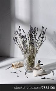 aromatherapy and organic concept - still life of dried lavender flowers in glass vase, craft sachet bag and rope dropping shadows on white surface. sachet bag, rope and lavender flowers in vase