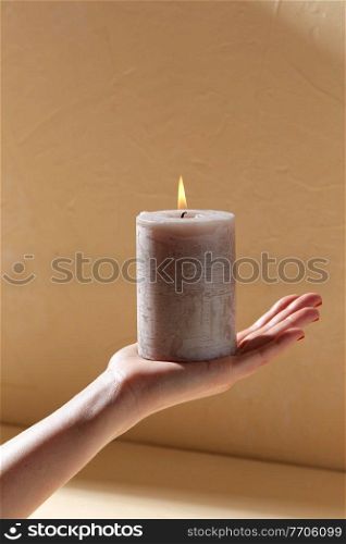 aromatherapy, coziness and people concept - female hand holding burning aroma candle on palm over beige background with shadows. hand holding burning aroma candle on palm