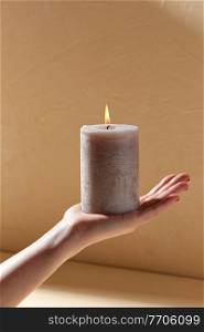 aromatherapy, coziness and people concept - female hand holding burning aroma candle on palm over beige background with shadows. hand holding burning aroma candle on palm