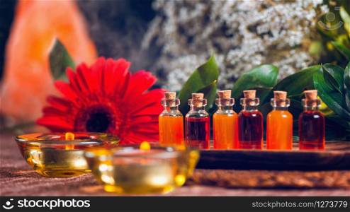 Aromatherapy relax concept with transparent bottles filled with red and orange essential oils on wooden board, red flower, candles and Himalaya salt lamp background.