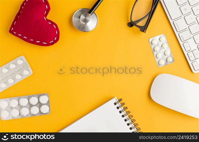 arrangement blister packed medicines stitched heart shape spiral notepad wireless keyboard mouse spectacles stethoscope yellow backdrop