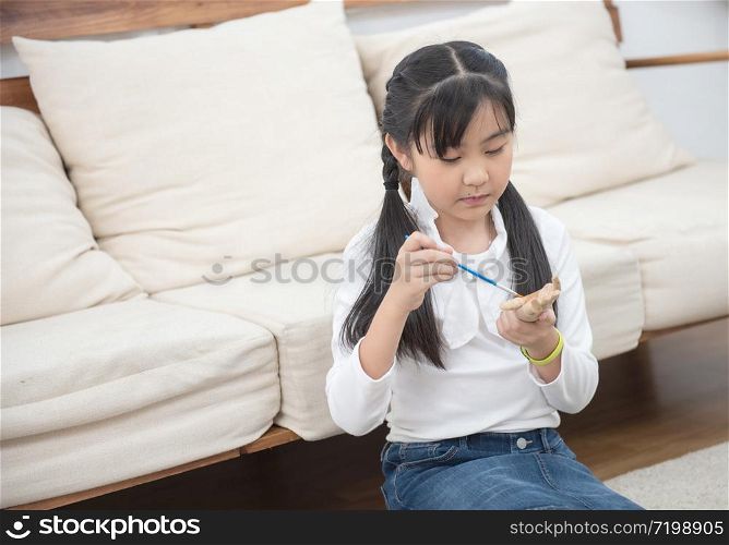 art asian girl student holding paint brush and learning paint on wood doll in living room at home on leisure weekend.