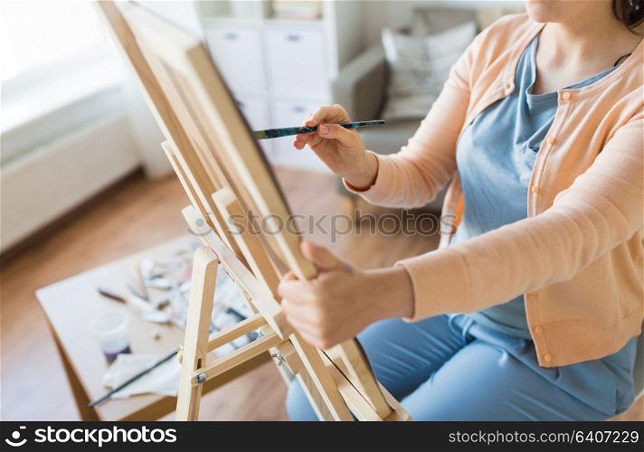 art, creativity and people concept - close up of artist with paint brush and easel painting at studio. artist with brush painting at art studio