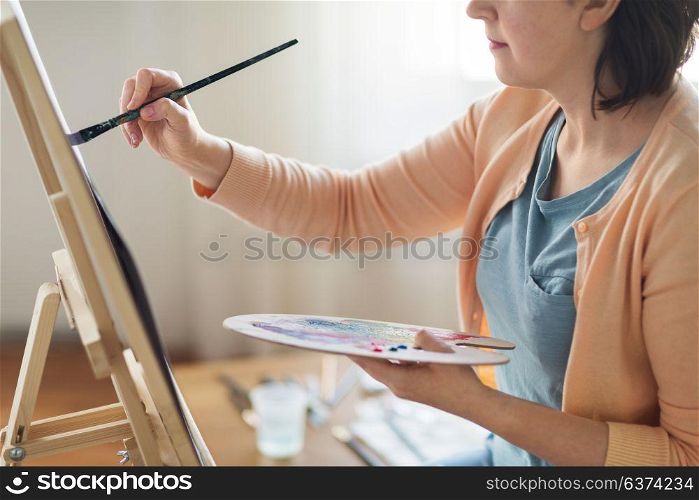 art, creativity and people concept - close up of artist with palette, brush and easel painting on paper at studio. artist with palette and brush painting at studio