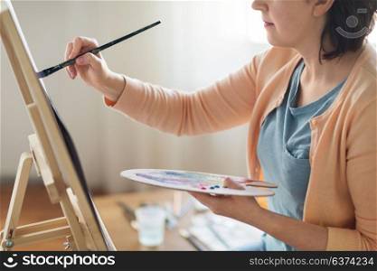 art, creativity and people concept - close up of artist with palette, brush and easel painting on paper at studio. artist with palette and brush painting at studio