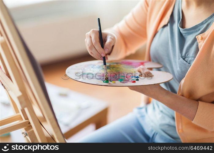 art, creativity and people concept - close up of artist with palette, paint brush and easel painting at studio. artist with palette and brush painting at studio