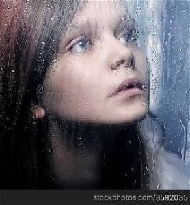 Art portrait of a beautiful young sadly girl standing at the window watching the rain