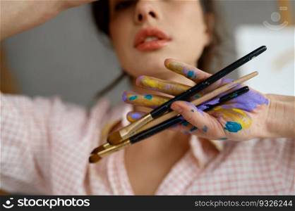 Art portrait of creative female artist with selective focus on dirty painted hands holding bunch of paintbrush. Cropped closeup shot of young painter. Art portrait of creative female artist showing hand with paintbrush