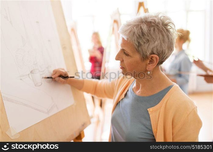 art school, creativity and people concept - happy senior woman artist with easel and pencil drawing picture at studio. woman artist with pencil drawing at art school
