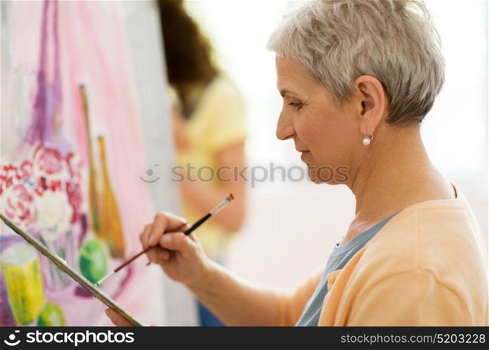 art school, creativity and people concept - happy senior woman artist with easel, paint brush and palette painting at studio. senior woman artist painting at art school