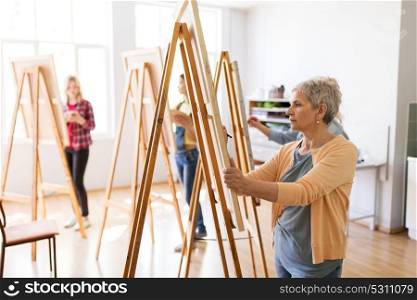 art school, creativity and people concept - happy senior woman artist with easel and pencil drawing picture at studio. artist with easel and pencil drawing at art school
