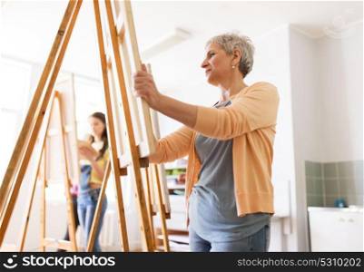 art school, creativity and people concept - happy senior woman artist with easel drawing at studio. woman artist with easel drawing at art school
