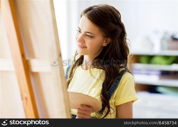 art school, creativity and people concept - happy smiling student girl or young woman artist with easel and palette painting at studio. student girl with easel painting at art school