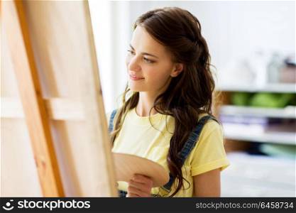art school, creativity and people concept - happy smiling student girl or young woman artist with easel and palette painting at studio. student girl with easel painting at art school