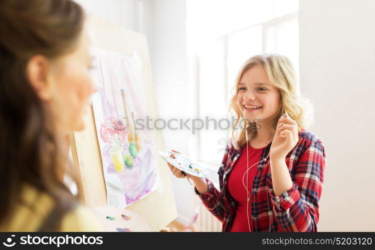 art school, creativity and people concept - happy smiling student girls or artists with earphones, easel and palette painting still life picture at studio. student girls with easel painting at art school