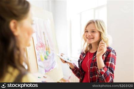 art school, creativity and people concept - happy smiling student girls or artists with earphones, easel and palette painting still life picture at studio. student girls with easel painting at art school