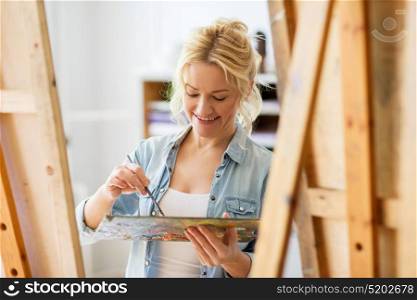 art school, creativity and people concept - happy smiling woman artist with easel, paint brush and palette painting at studio. happy woman with easel painting at art school