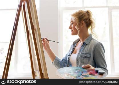art school, creativity and people concept - happy smiling woman artist with easel, paint brush and palette painting at studio. woman artist with easel painting at art studio
