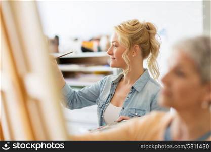 art school, creativity and people concept - happy smiling woman artist with easel, paint brush and palette painting at studio. woman artist with brush painting at art school