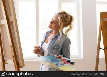 art school, creativity and people concept - happy woman artist with easel, paint brush and palette painting at studio. woman artist with easel painting at art school