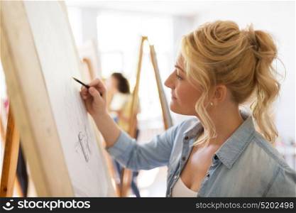 art school, creativity and people concept - happy woman artist with easel and pencil drawing picture at studio. woman artist with pencil drawing at art school