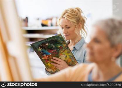 art school, creativity and people concept - happy woman artist with paint palette painting at studio. woman artist with palette painting at art school