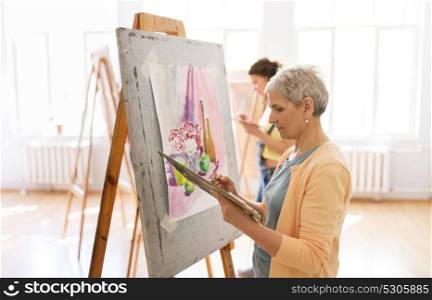 art school, creativity and people concept - senior woman artist with easel, paint brush and palette painting at studio. woman artist with easel painting at art school